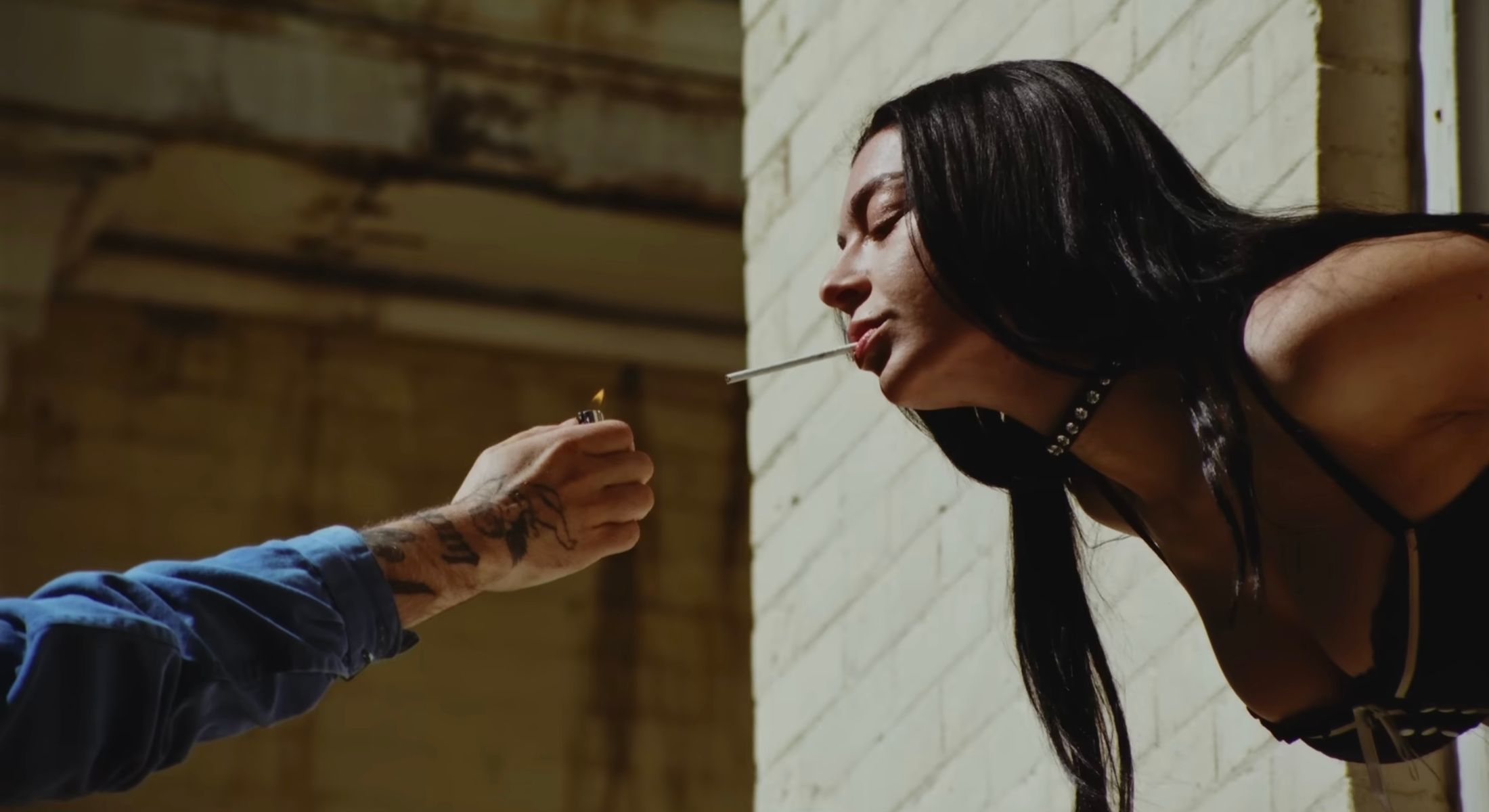 On the right of the frame, a young woman with long black hair leans out a window towards a person in a blue shirt holding a lighter for the cigarette in her mouth. Behind them is a white brick wall.
