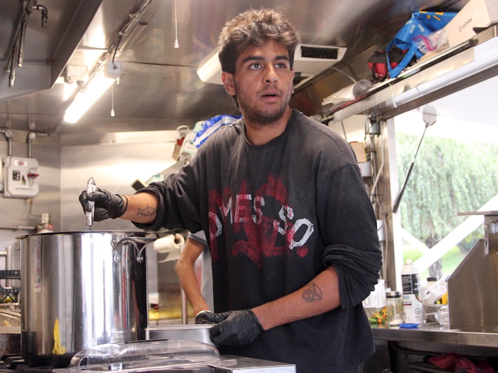 A young man stirs a large pot in a small mobile kitchen while looking beyond the camera.