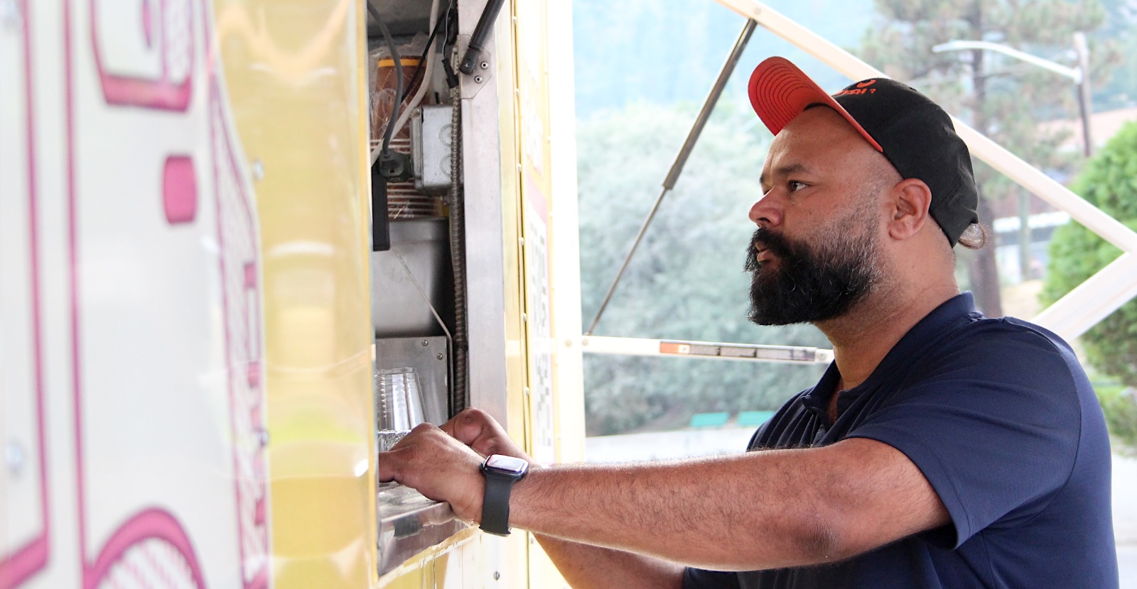 A man with a large beard and wearing a hat leans on the window of a food truck.