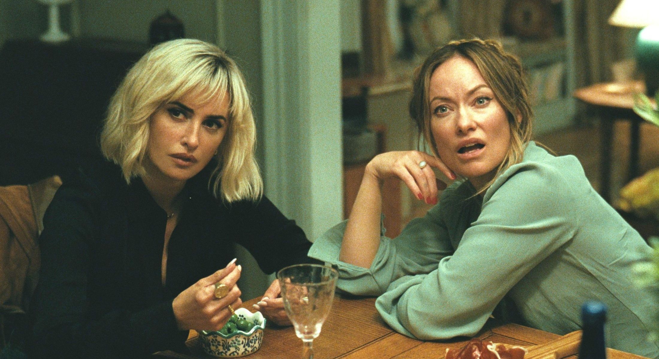 Penelope Cruz as Pina and Olivia Wilde as Angela sit at a table looking shocked in a still of 'The Invite.' 