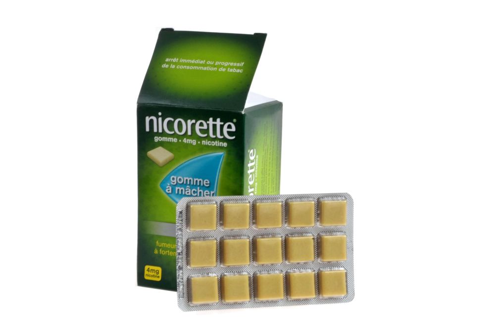 A green package of Nicorette brand nicotine gum stands against a white background. Its interior blister pack of gum stands beside the box. The gum tablets are a beige colour.