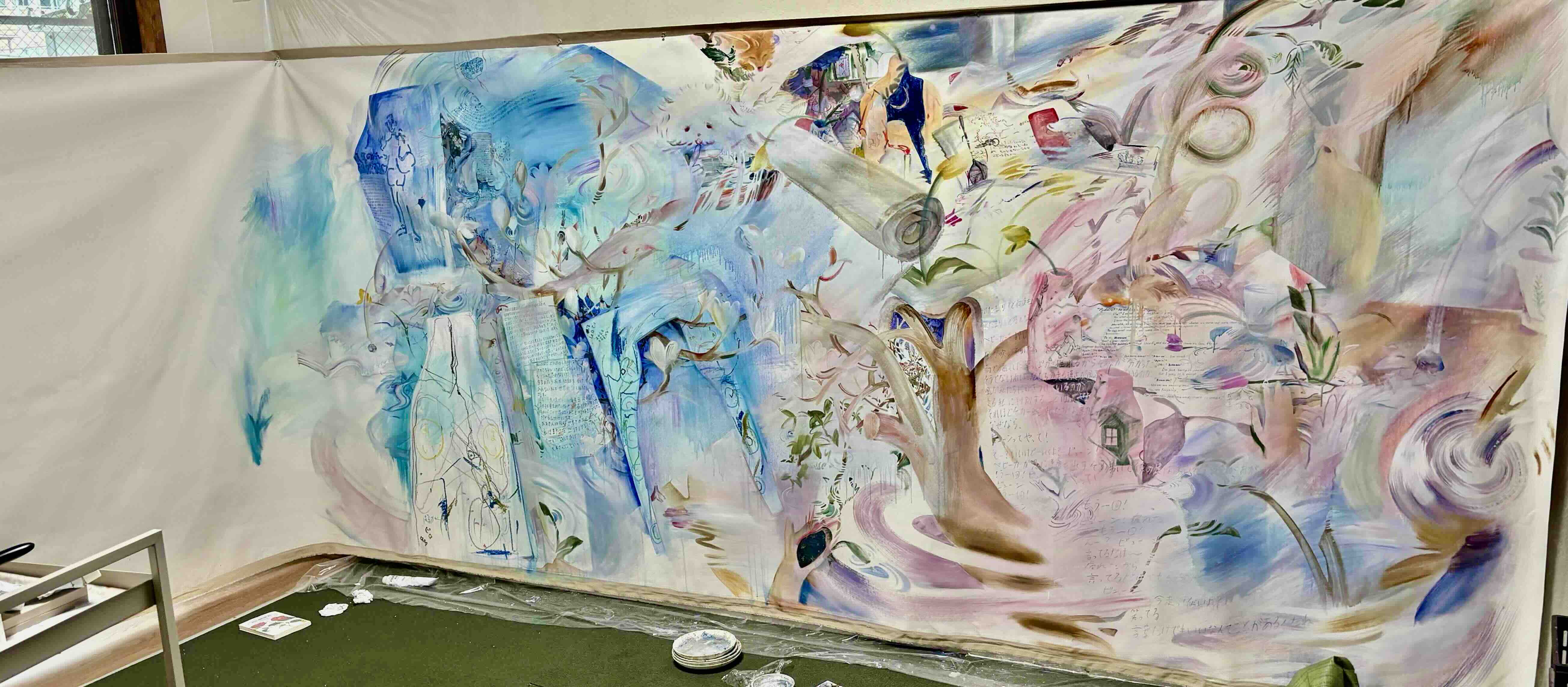 A wide-format painting in light tones features branches, flowers and other abstract shapes across a wall-length canvas in a studio setting.