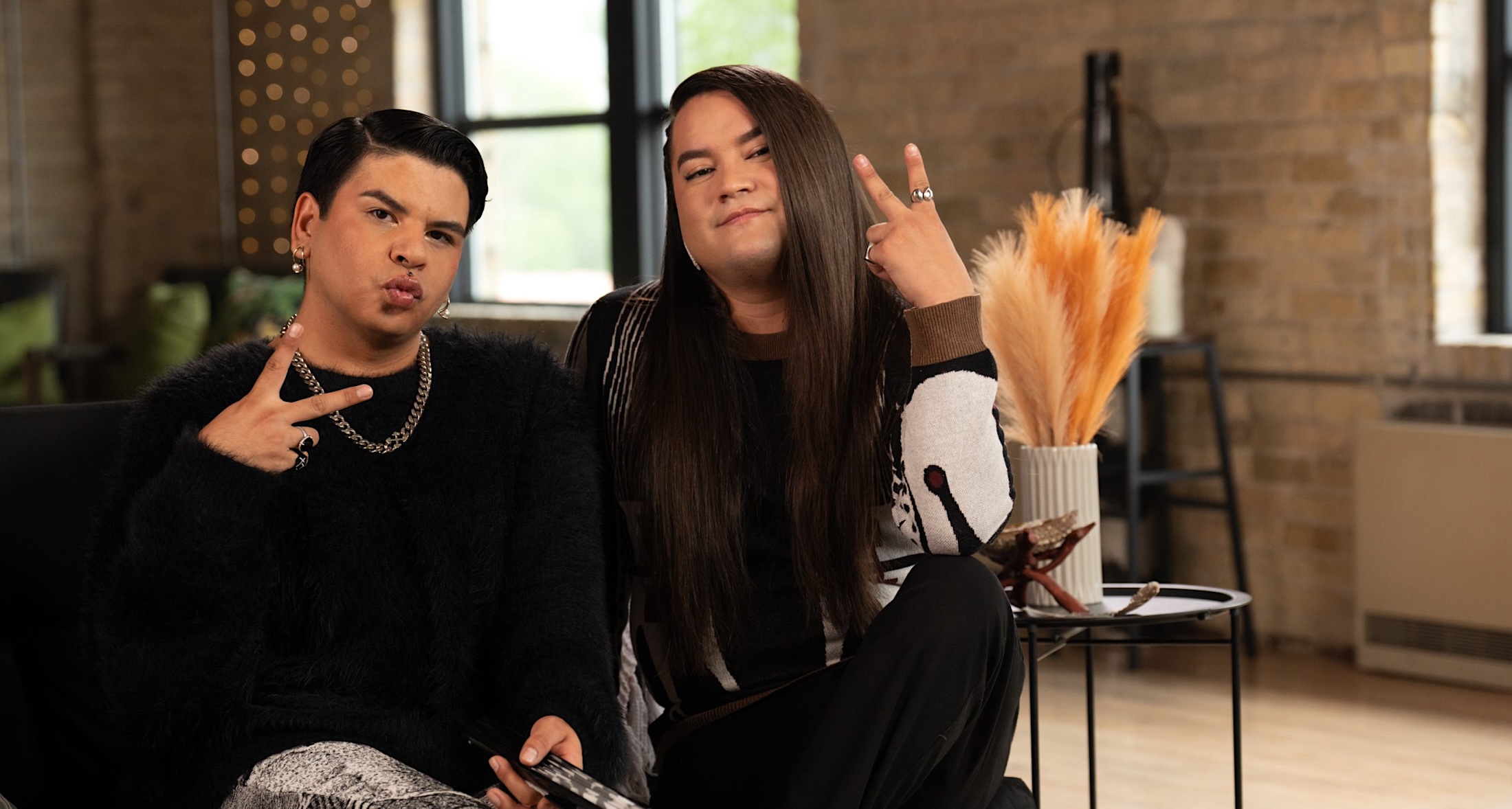 Scott Wabano, left, is seated on a sofa with Kairyn Potts, right. They are posing and holding two fingers up in a peace sign. They are wearing dark clothing. Wabano has short dark wavy hair and Potts has long brown hair.