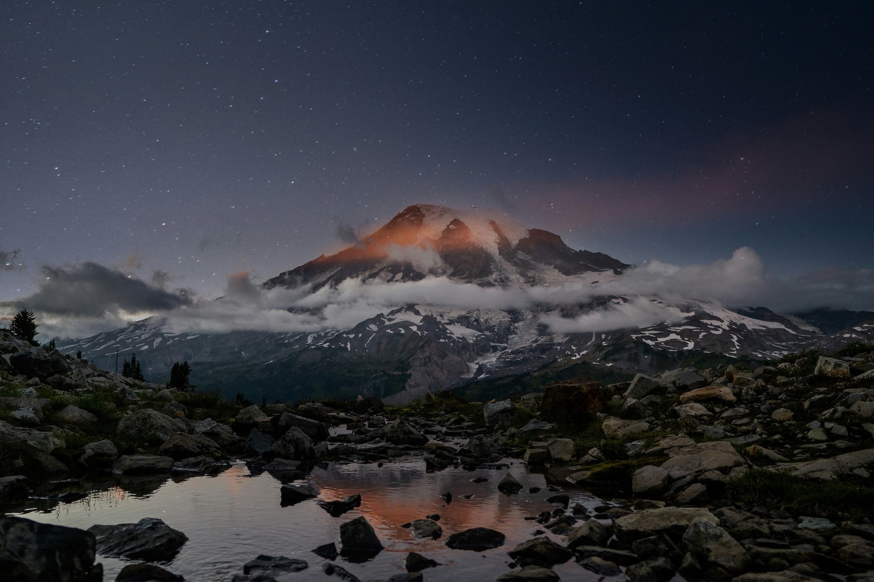 A panoramic photo of a mountain range at dusk. A bank of clouds rings its midsection. In the foreground is a rocky body of water. The sky is dark grey with stars.