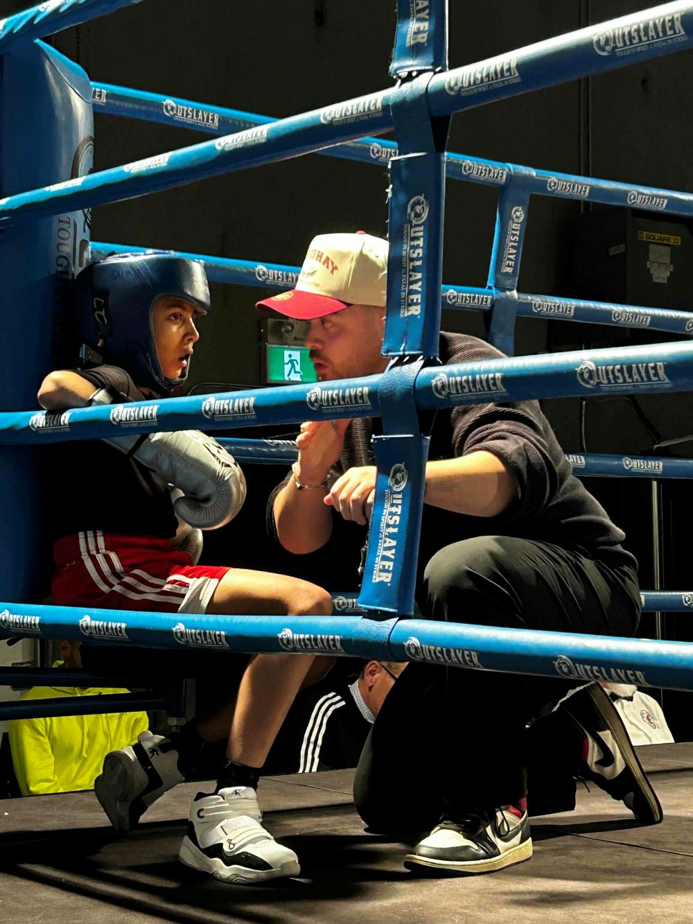 Sol Diana, right, wears a white and red ball cap and dark clothes. He is crouching on one knee, speaking to a child in a blue boxing helmet, red shorts and silver gloves resting against the ropes in the corner of a boxing ring.