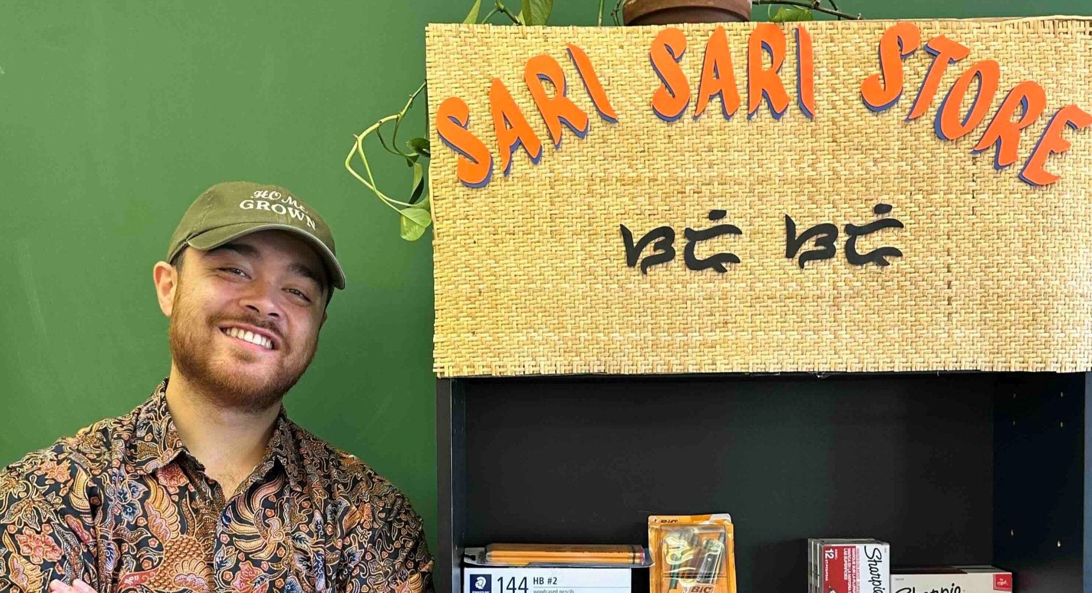 Sol Diana wears a ball cap, a short sleeved button-down shirt with a paisley pattern, and navy trousers. His arms are crossed and he is smiling. He stands next to a bookshelf full of school supplies with a handmade sign that says &ldquo;Sari Sari Store&rdquo; above it.