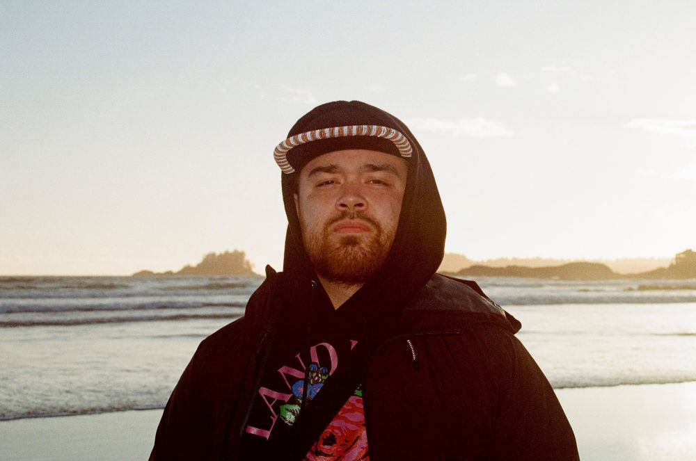 Sol Diana is wearing a black hoodie over a ball cap. The hoodie has a floral pattern featuring the words &ldquo;Land Back.&rdquo; He is standing on a beach at sunset, looking directly at the camera.