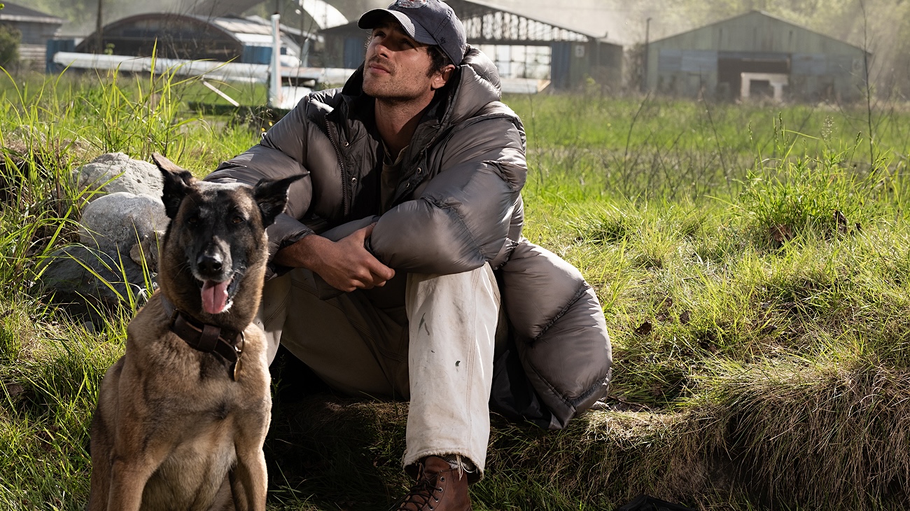 A man with dark hair and a dark ball cap sits in green grass, looking up towards the left of the frame. He wears a long black puffer jacket and khakis. A German shepherd sits with him to the left of the frame.