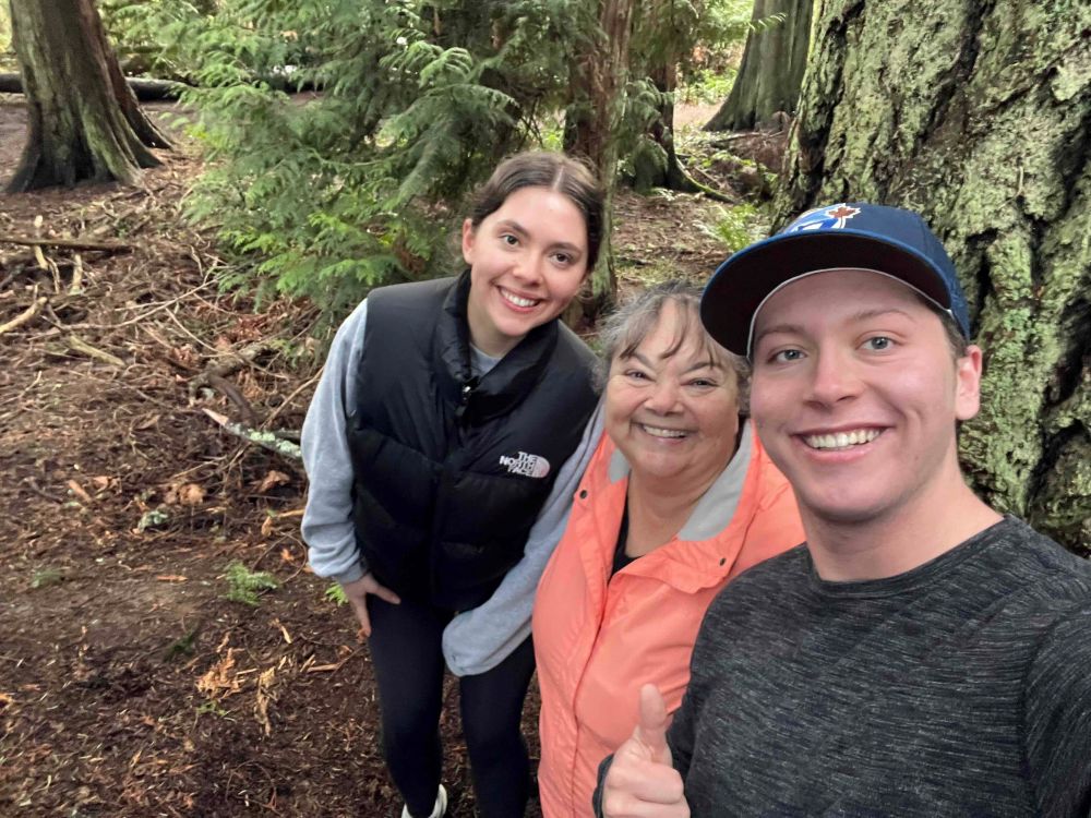 Monique Gray Smith, centre, is wearing a peach-coloured rain jacket. She is smiling at the camera and flanked by two young adults in grey, black and white streetwear. They are standing next to a tree in a forest.