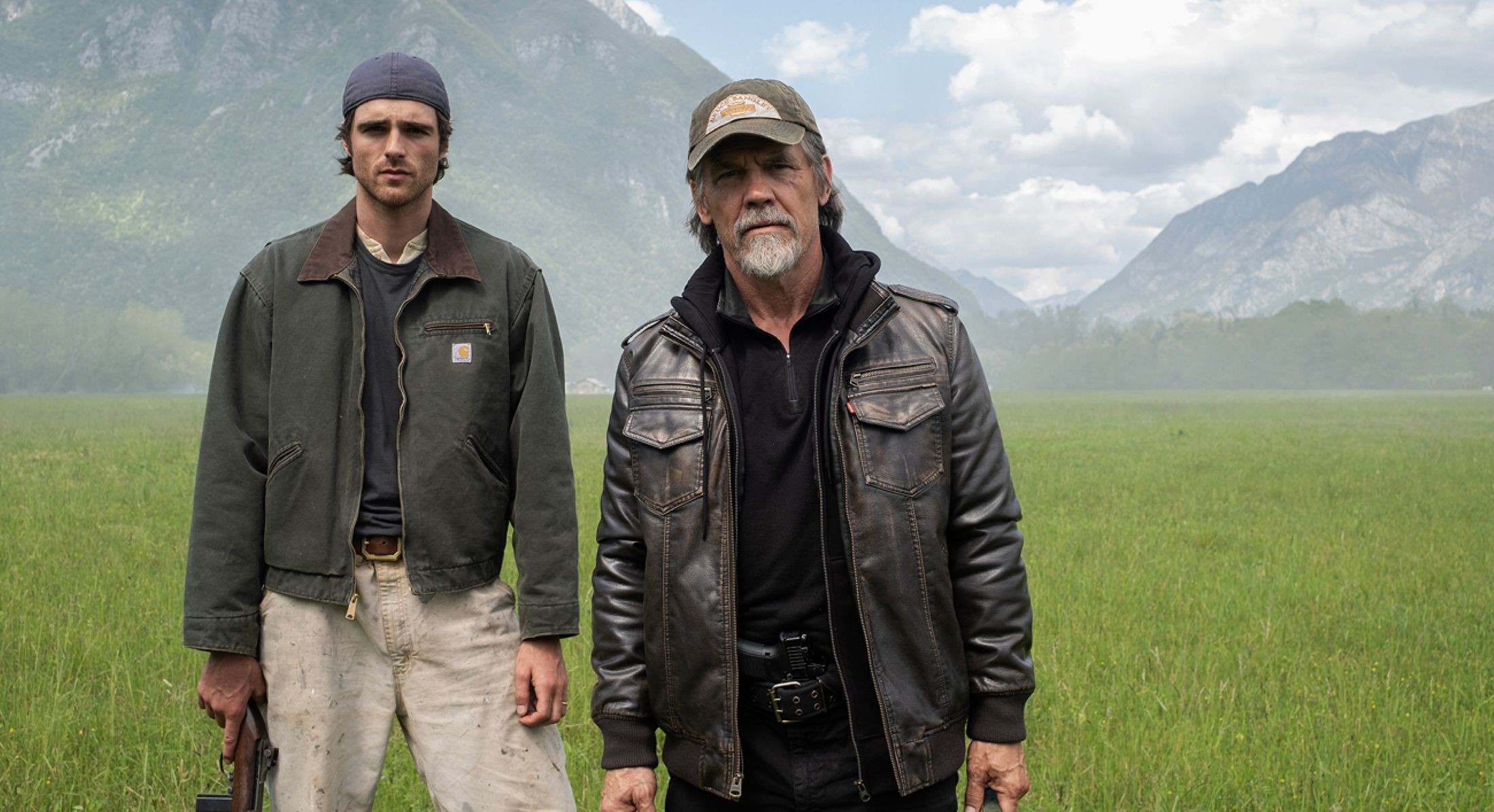 Two actors stand on a movie set in a grassy meadow against a mountainous backdrop. Jacob Elordi, left, is wearing a backwards ball cap, an olive-green chore coat and khakis. Josh Brolin, right, is an older man in a dark ball cap and leather jacket over a black outfit.