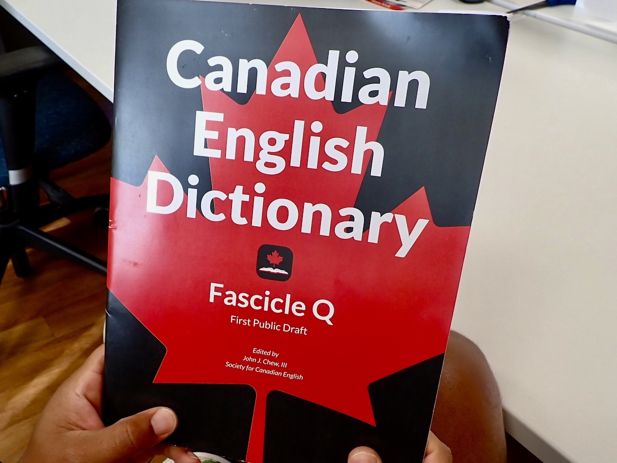 Two hands hold up a navy blue book with a red maple leaf under white text that reads, &lsquo;Canadian English Dictionary. Fascicle Q. First public draft.&rsquo;