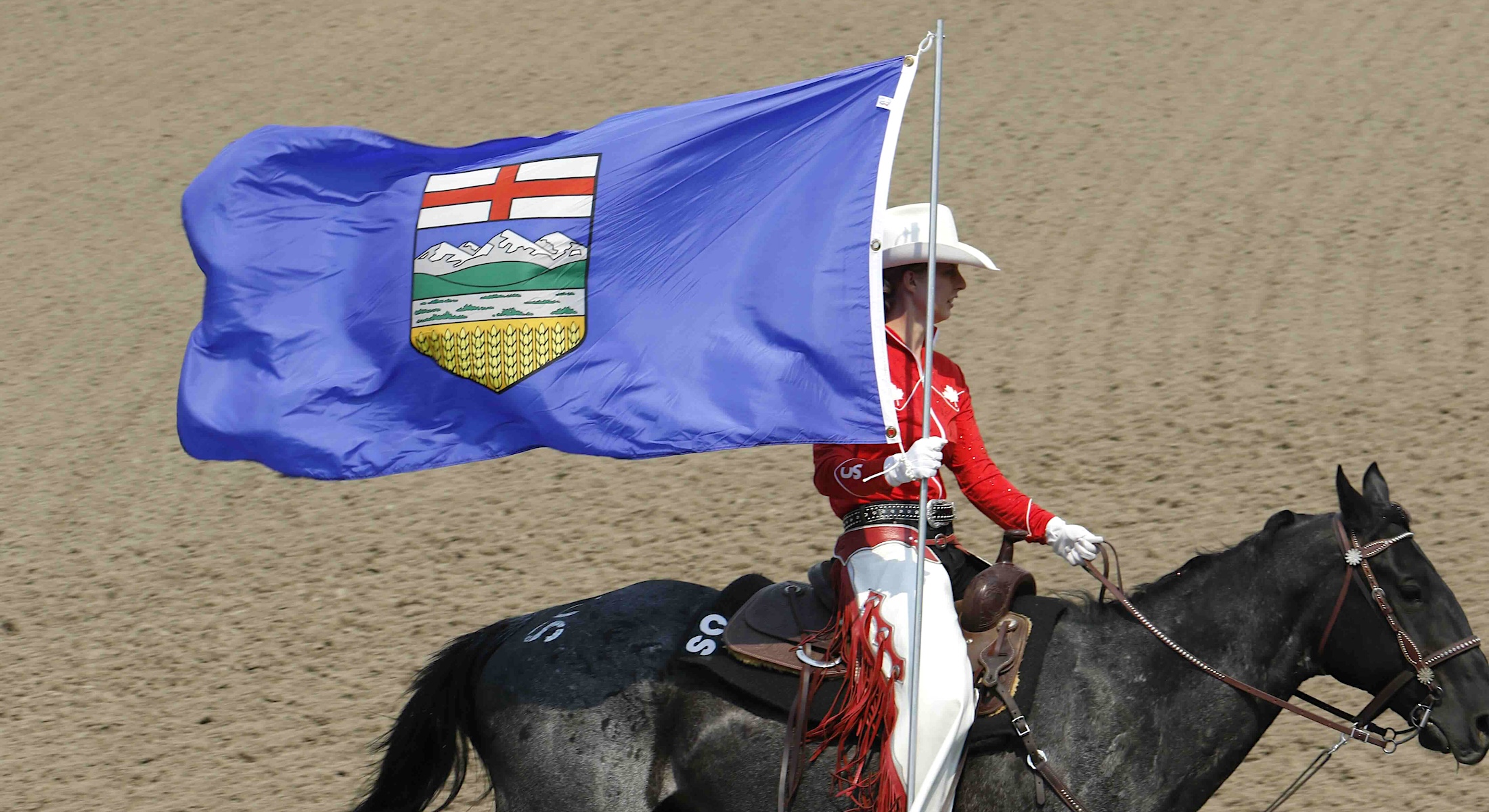 A person in a red rodeo uniform rides a black horse while carrying the blue Alberta provincial flag across the dirt floor of an arena.