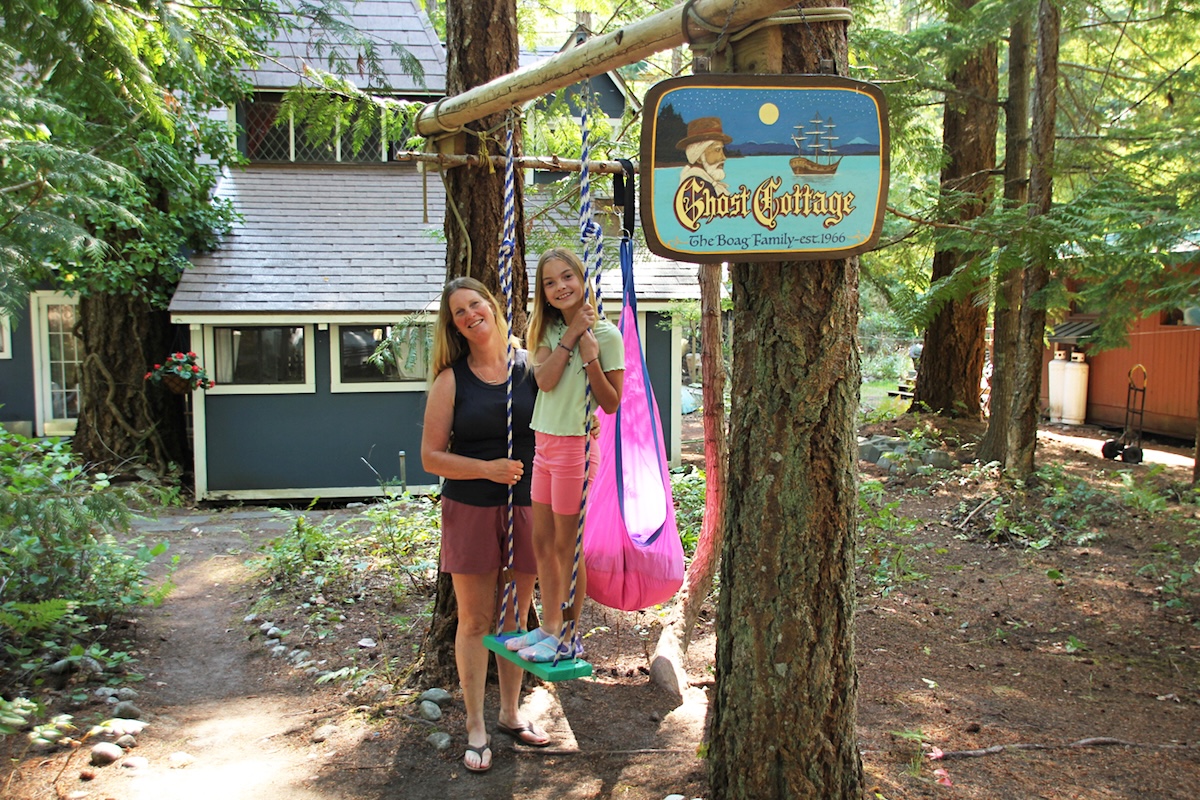 A young girl and her mom hang out by a swing hanging from a tree. The tree has a sign: &lsquo;Ghost Cottage. The Boag Family. Est. 1966.&rsquo;