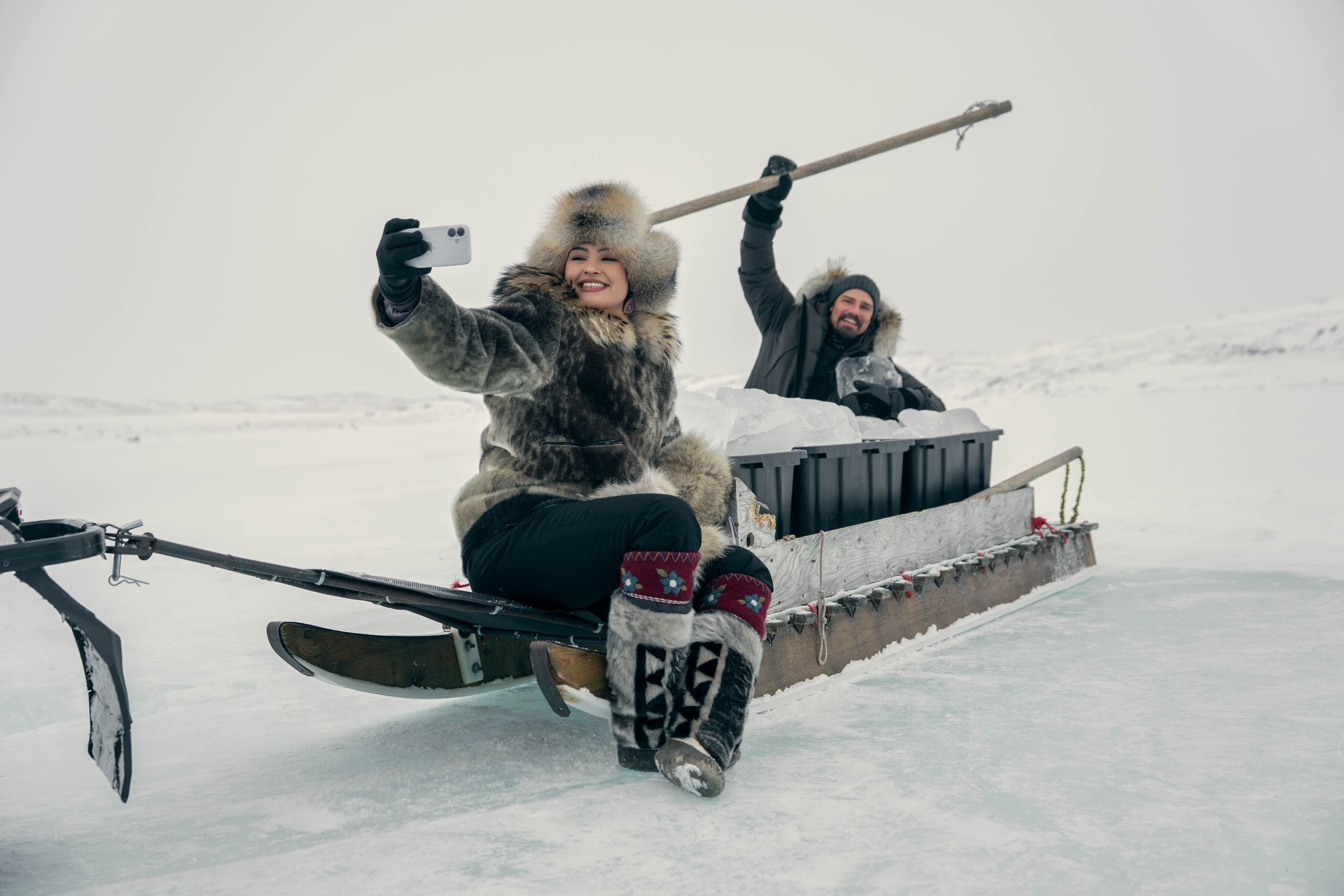 A woman in a traditional Inuk winter fur outfit sits on the front of a dogsled on a snowy field, pausing to take a selfie of herself and the man behind her, in a grey winter jacket, holding a long wooden tool.