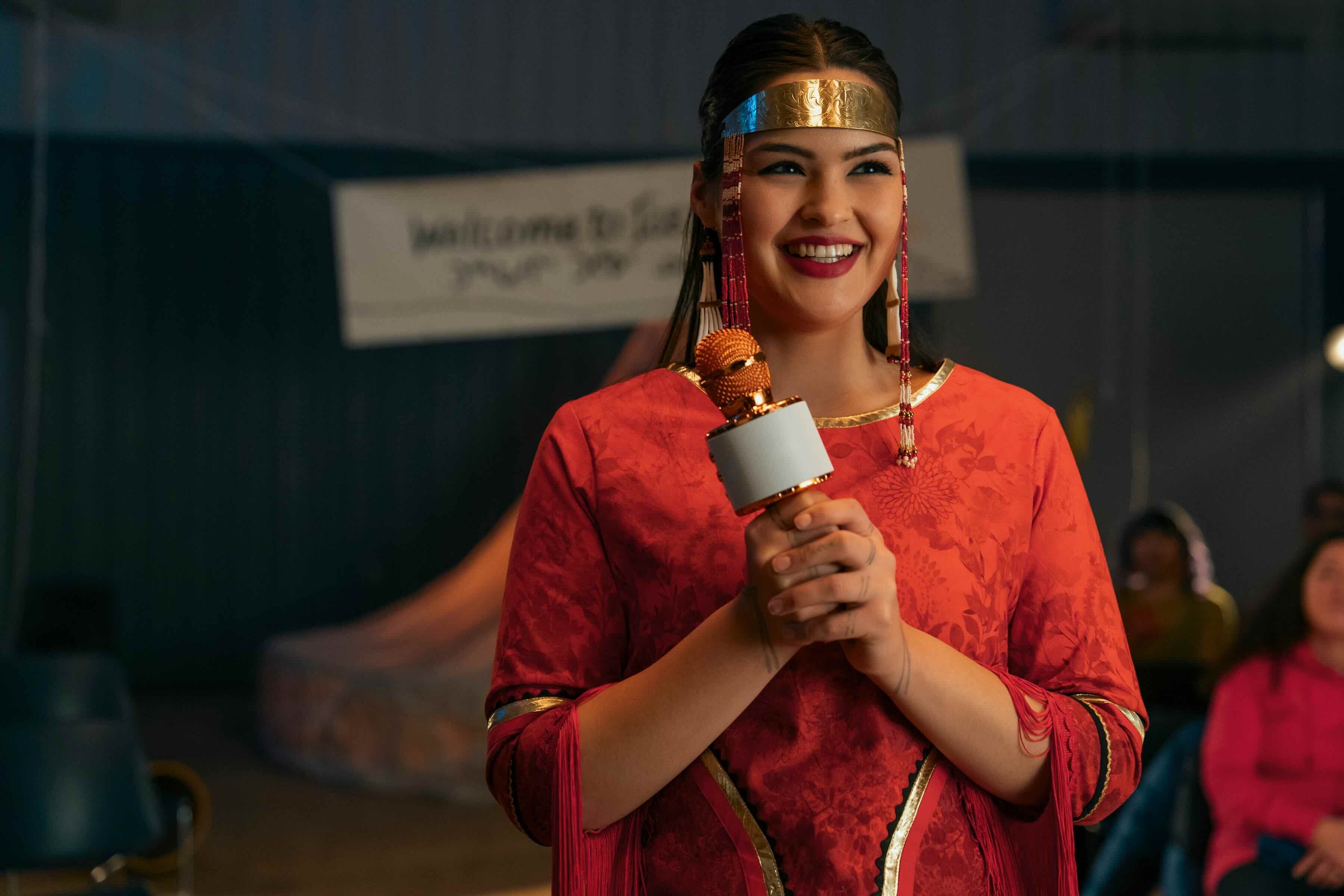 Actor Anna Lambe is holding a microphone and smiling. She is wearing a a traditional red dress and a gold headband lined with beadwork.