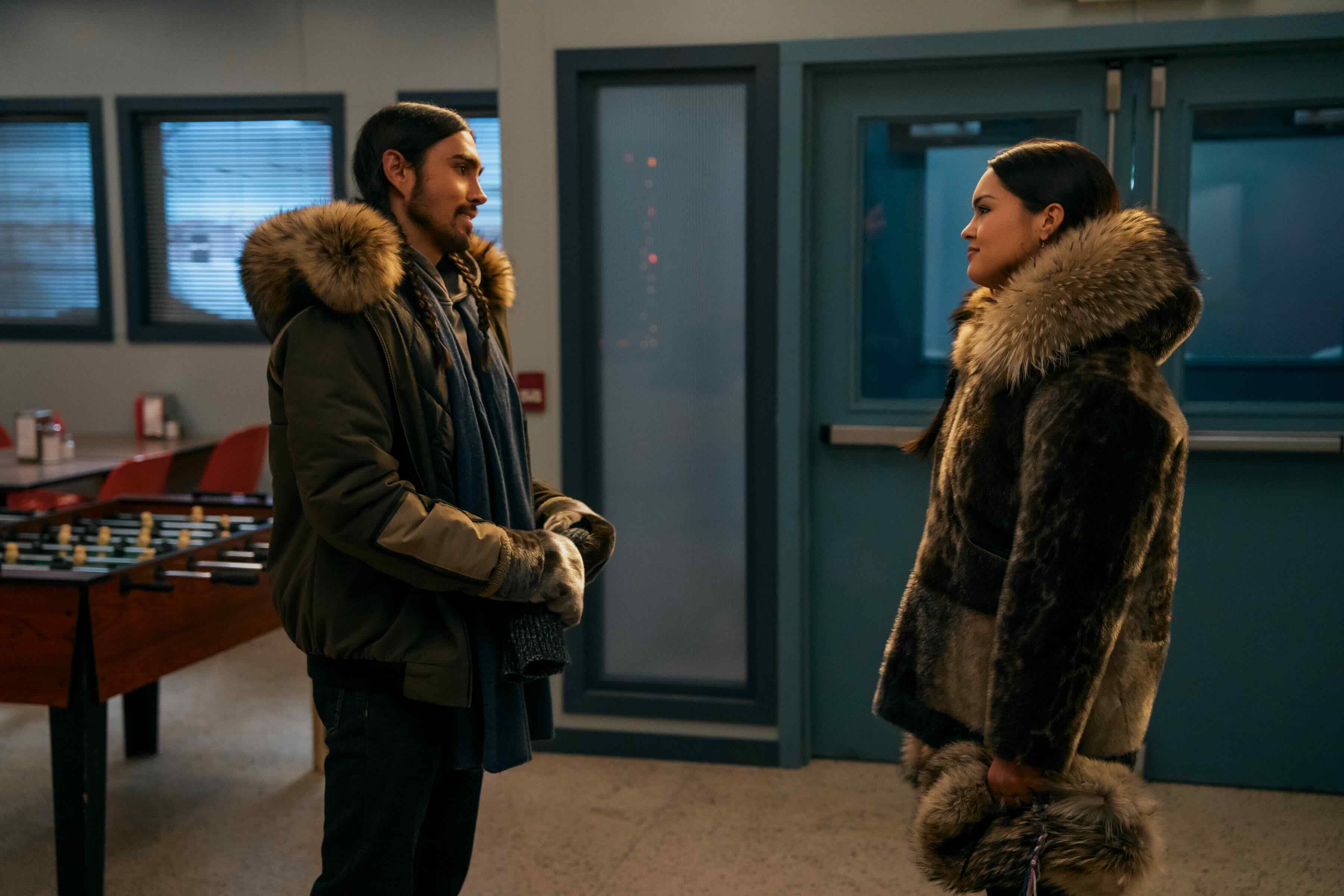 Two actors, a man on the left and a woman on the right, stand in an indoor setting with a foosball table in the background. They are wearing winter jackets lined with fur. Both have long dark hair, and they are looking at each other.