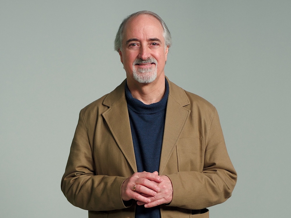 Max Cameron is wearing a tan blazer over a navy shirt. He is standing against a grey studio background, smiling. He has a beard and grey hair.