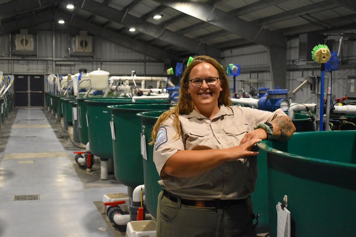 A woman with light skin tone and long light brown hair leans on a sturgeon tank in a large building.