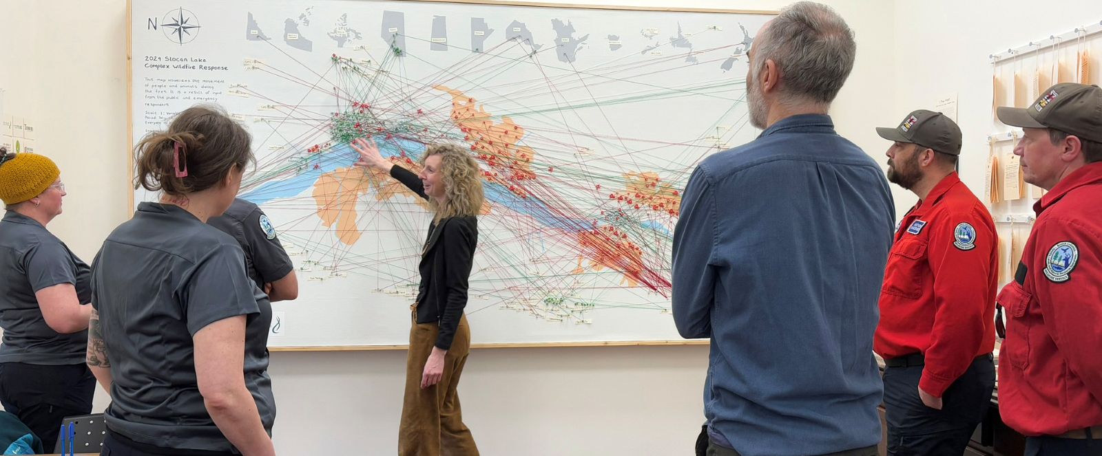 A woman explains a map and an array of red and green pins and strings to six visitors. Shoulder badges show that at least three are members of the BC Wildfire Service.