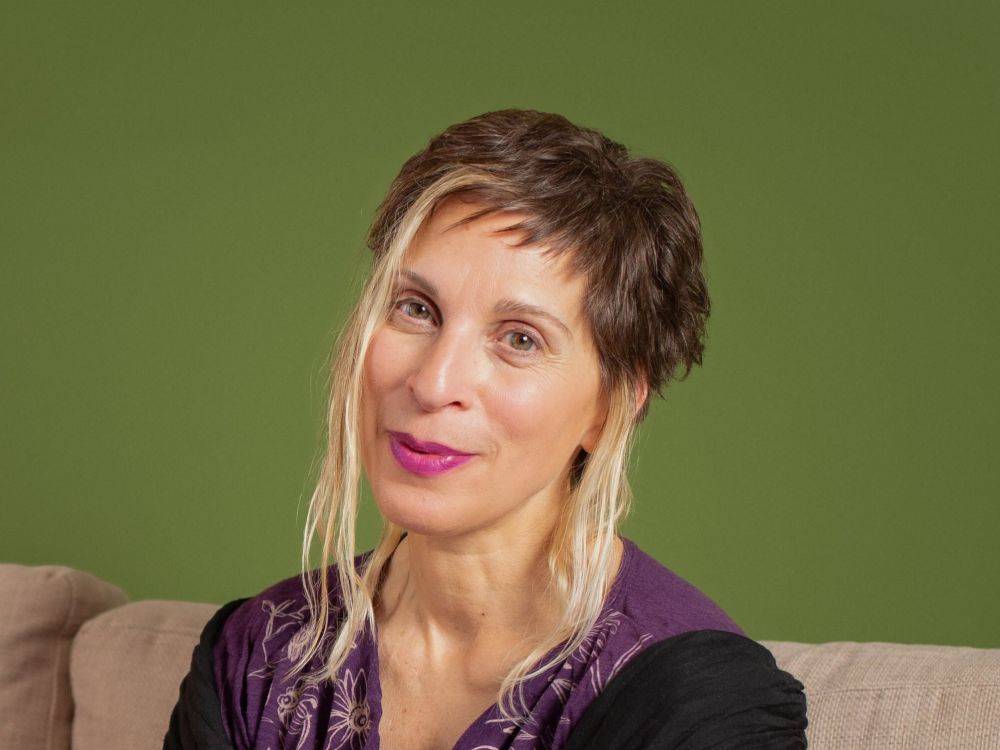 Leilani Farha is seated on a beige sofa against a green background. She wears a purple patterned top under a black cardigan.