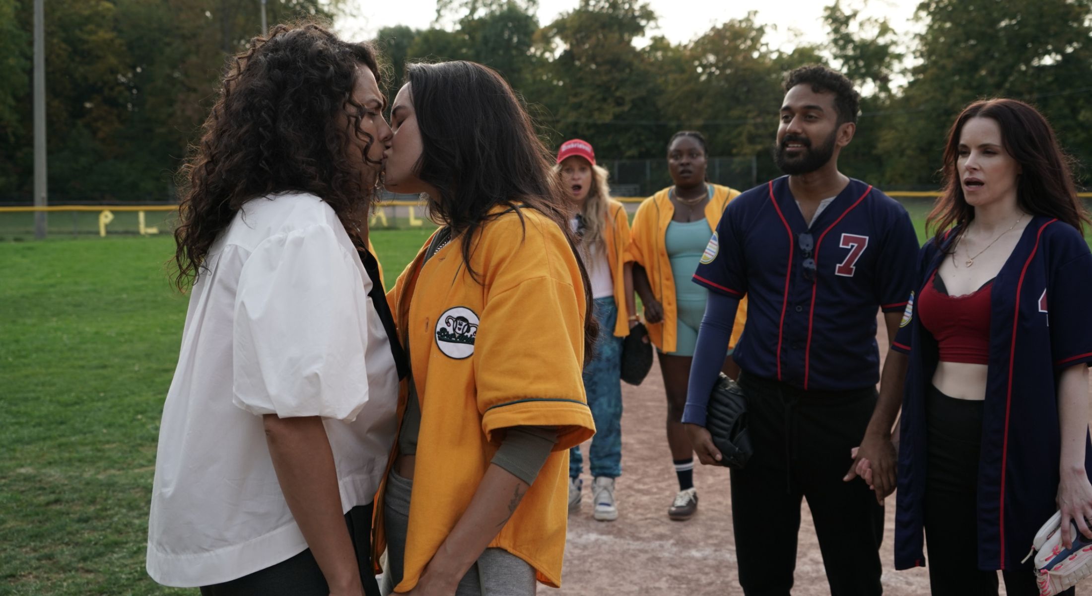Actors in blue and yellow baseball uniforms stand outside on a baseball diamond. Two women, one in a white shirt and one in a yellow baseball uniform, are standing to the left of the frame, kissing, while others look on in surprise.