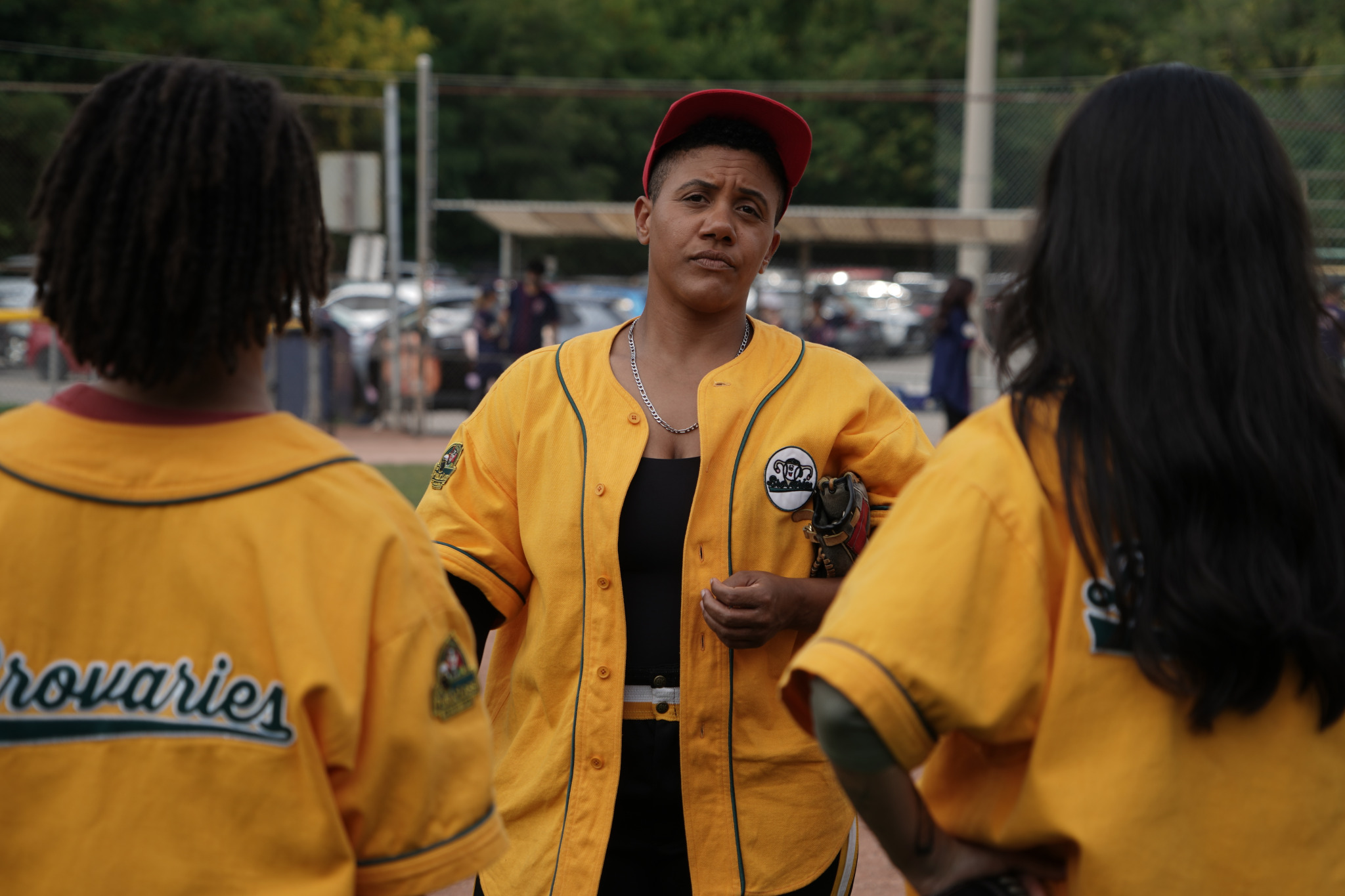 A person in a yellow baseball uniform stands with an exasperated expression on their face. They are facing two players in similar uniforms, their backs turned to the camera.