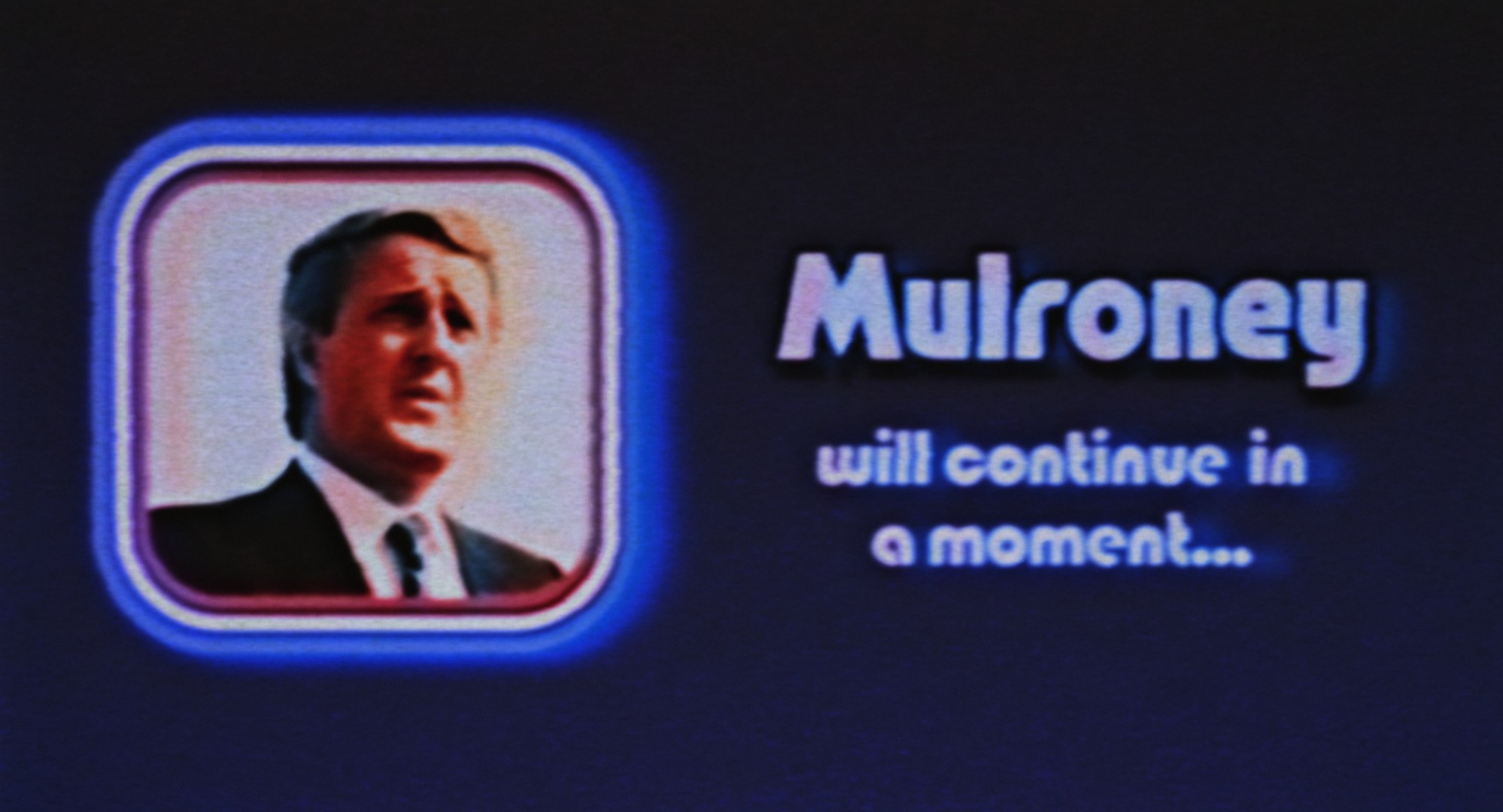 A pixelated screen features a square with a man in a suit shot from the chest up. Next to it in blue text reads &ldquo;Mulroney will continue in a moment&rdquo; against a black background.