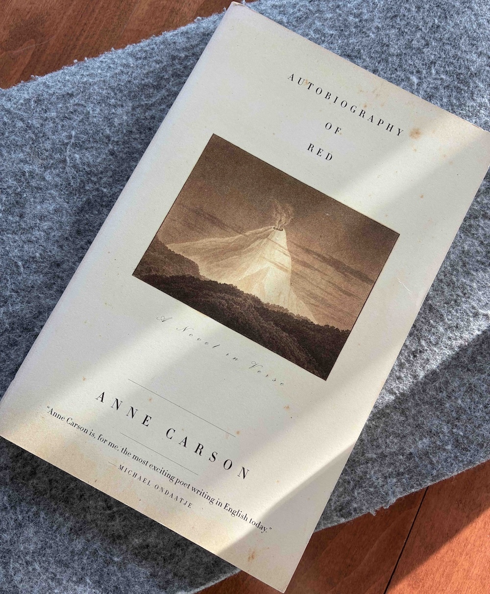 A photo of the book cover image of <i>The Autobiography of Red</i> in sunlight against a grey soft background and wood tabletop. The book cover is off-white and features a small illustration of a red volcano.