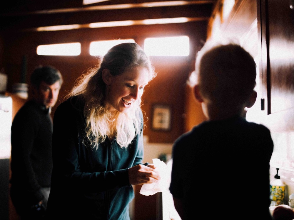 Elizabeth Holmes is standing at a kitchen counter, looking down at an object in her hands and smiling. The back of a toddler is in the foreground, facing Holmes, who has long blond hair. In the background, Nathan Fielder stands watching to the left of the frame. He has short thick dark hair and is wearing dark clothing.