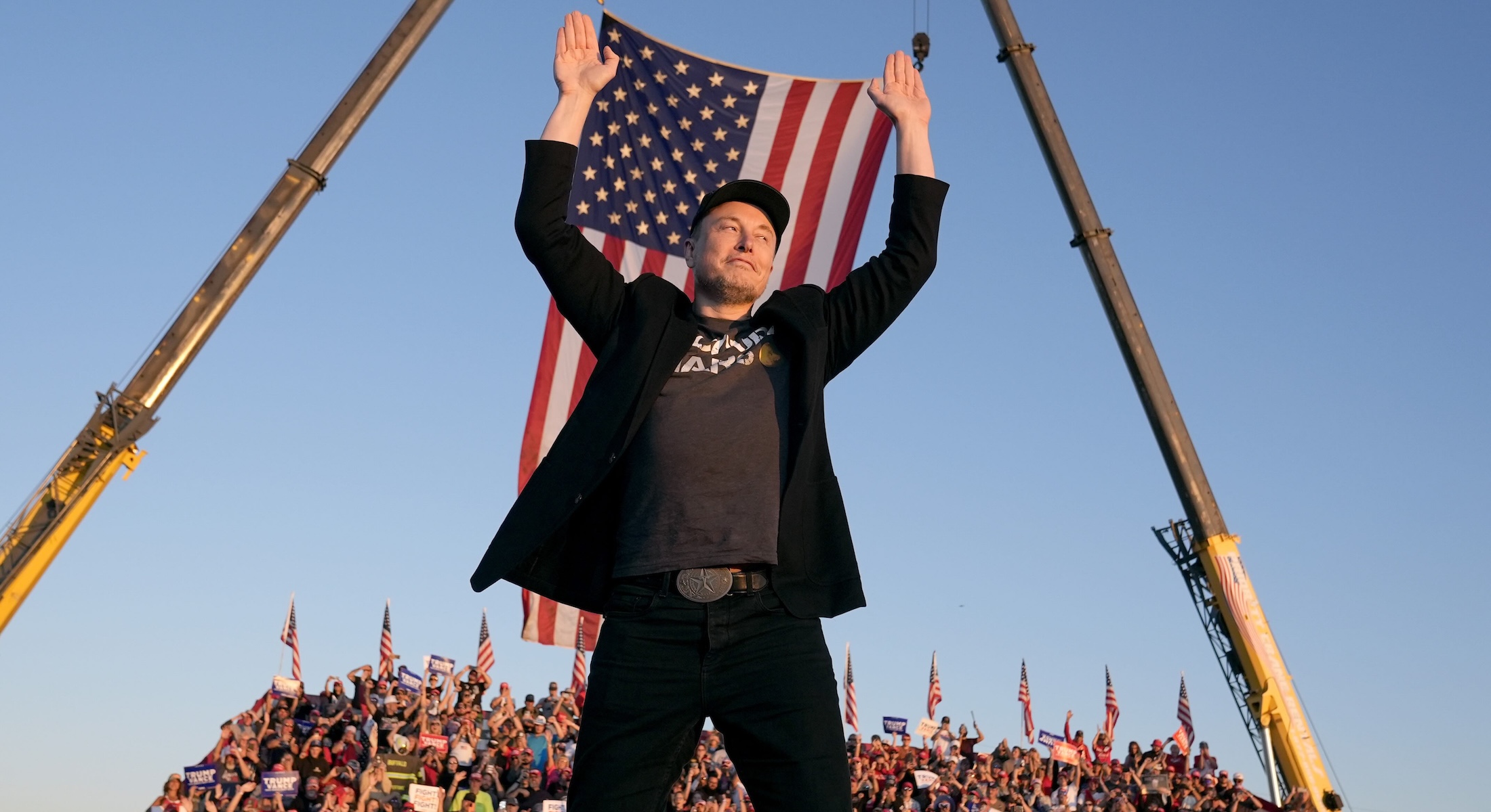 Elon Musk wears a black ball cap, black jeans, a dark grey T-shirt and a black blazer. He stands on an outdoor stage, arms raised against the backdrop of an American flag, a grandstand full of people and a sky at sunset.