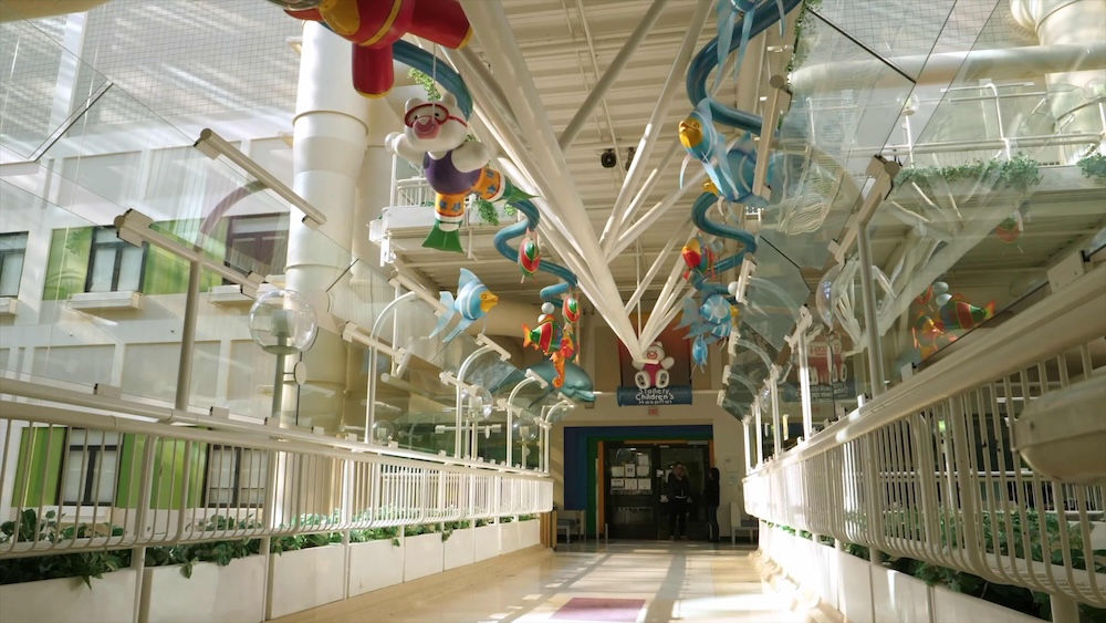 An indoor hospital walkway features white railings, a white lamppost and a colourful ceiling installation of cartoon animals swimming in the sea.