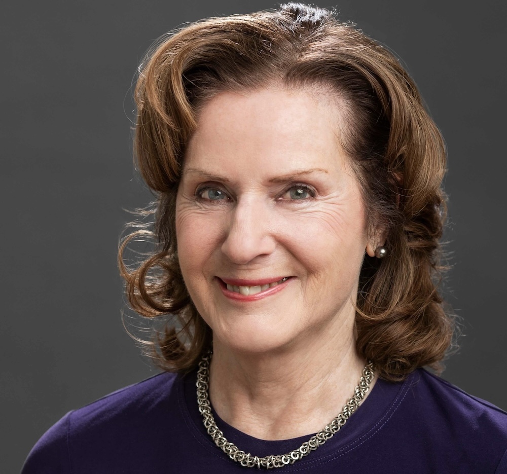 A portrait of Carol Off, who has shoulder-length curly brown hair. She wears a navy sweater with a silver necklace.