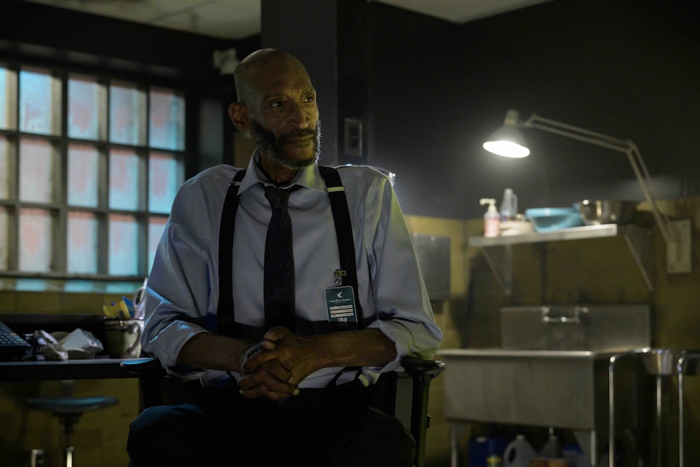 Actor Tony Todd wears suspenders over a light blue button-down and dark tie. He is seated in a film set in a dim laboratory space. He is bald with a dark beard.