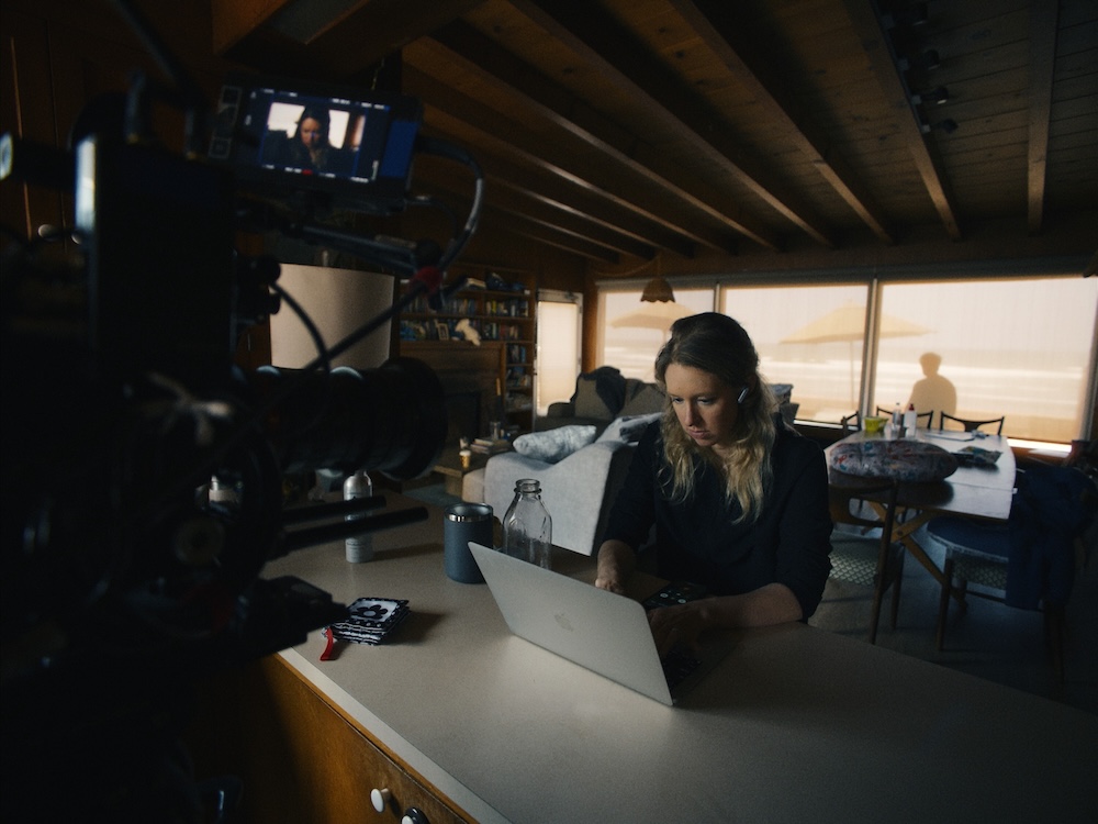 Elizabeth Holmes is seated at a kitchen counter, working on a silver Macbook Air laptop. She has long wavy blond hair and she is wearing a white AirPod in one ear while she types on the computer. Behind her is a large living room and bay windows that look out to the water. An array of cameras and equipment darken the left of the frame.