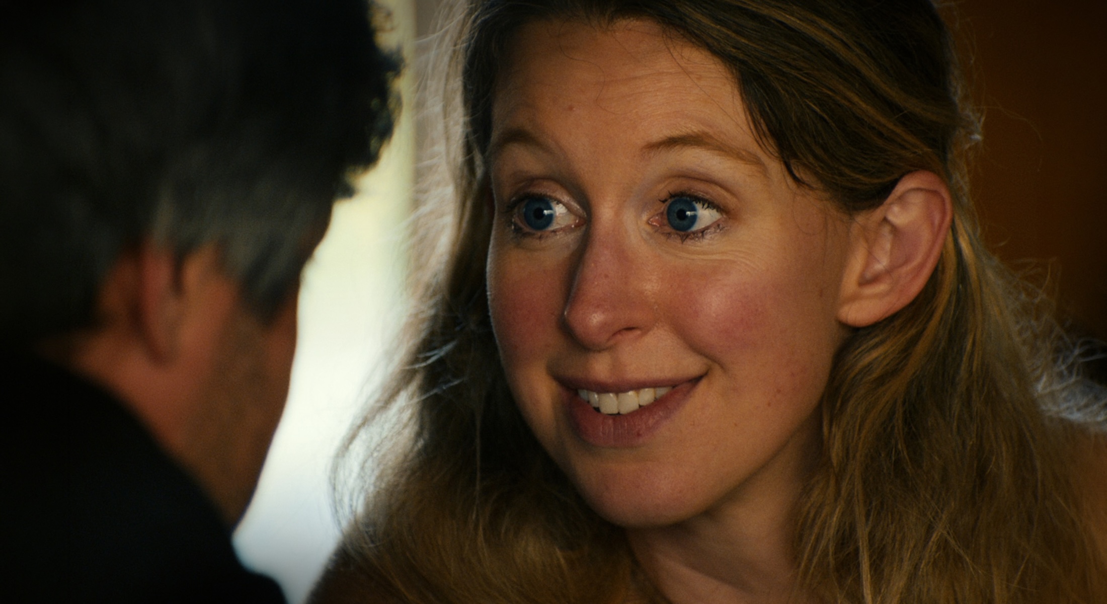 Elizabeth Holmes has wide blue eyes. She is leaning towards the person speaking to her, who has dark hair and whose back is turned to the camera. She has long blond hair and she is wearing a beige sweater.