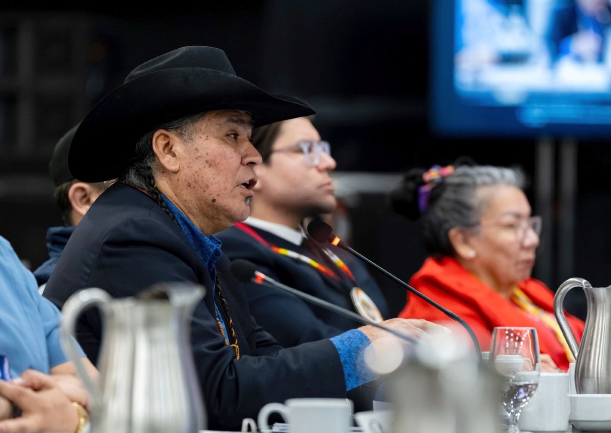 A First Nations man with a cowboy hat speaks into a microphone while seated at a long table with other leaders.