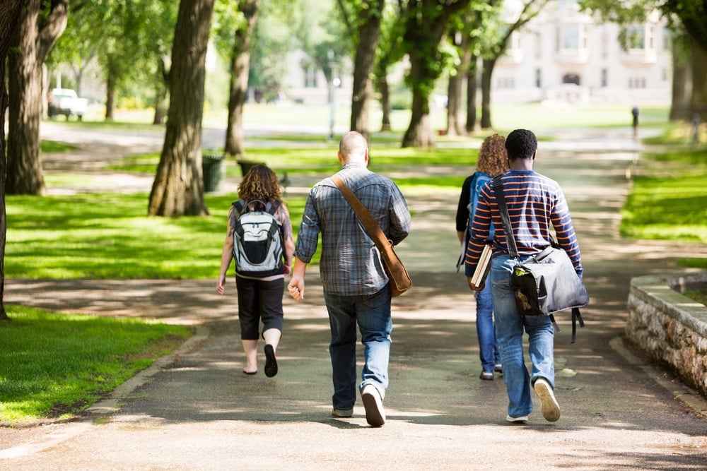 Students wearing messenger bags and backpacks walk down a path on a university campus.