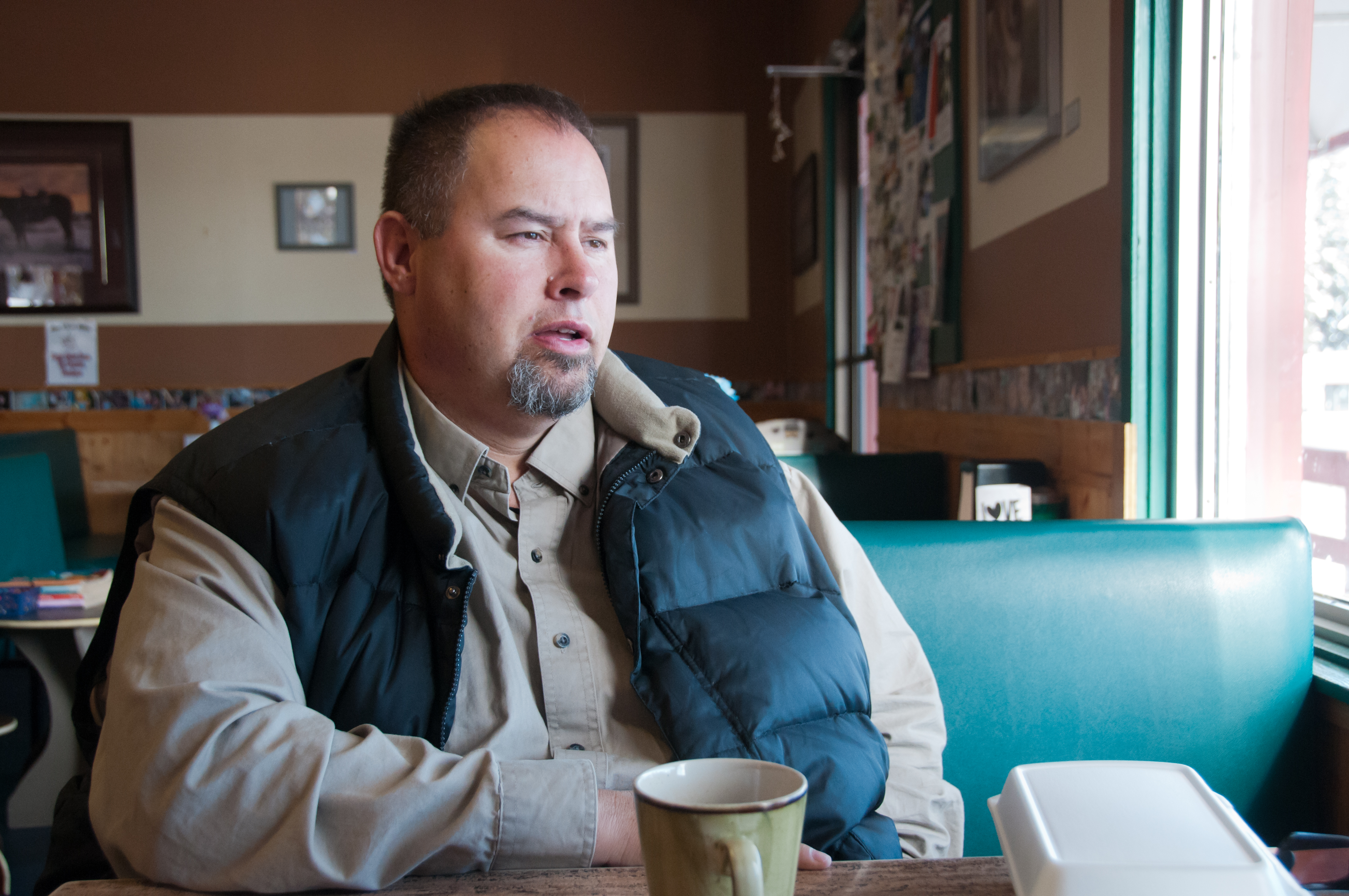 A man sits at a booth in a diner on a turquoise banquette. He has short dark hair, a goatee and is wearing a light brown button-down shirt and a black down vest.
