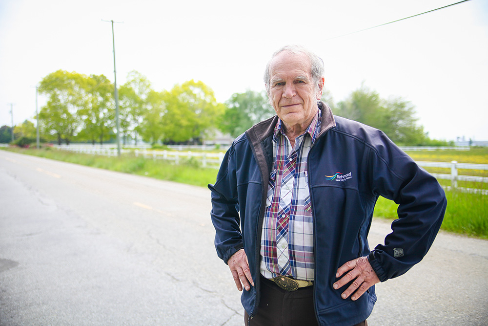 A senior man stands with his hands on his waist. He is wearing a navy jacket with the City of Richmond logo, the vague shape of a heron made out of colourful ribbons. He is on a paved road in the middle of farmland.