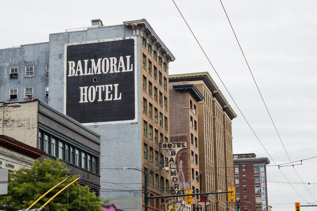 A view of the Balmoral Hotel on East Hastings Street in Vancouver’s Downtown Eastside. The Balmoral has its name prominently painted in large white text inside a black square on the side of the building, which is painted light grey. To the right, a curvy neon sign that reads “Balmoral” hangs off front the side of the tall multi-storey building, painted brown. Other tall brown buildings are beside it and bus cables above the street are visible in the shot. The sky is grey. 