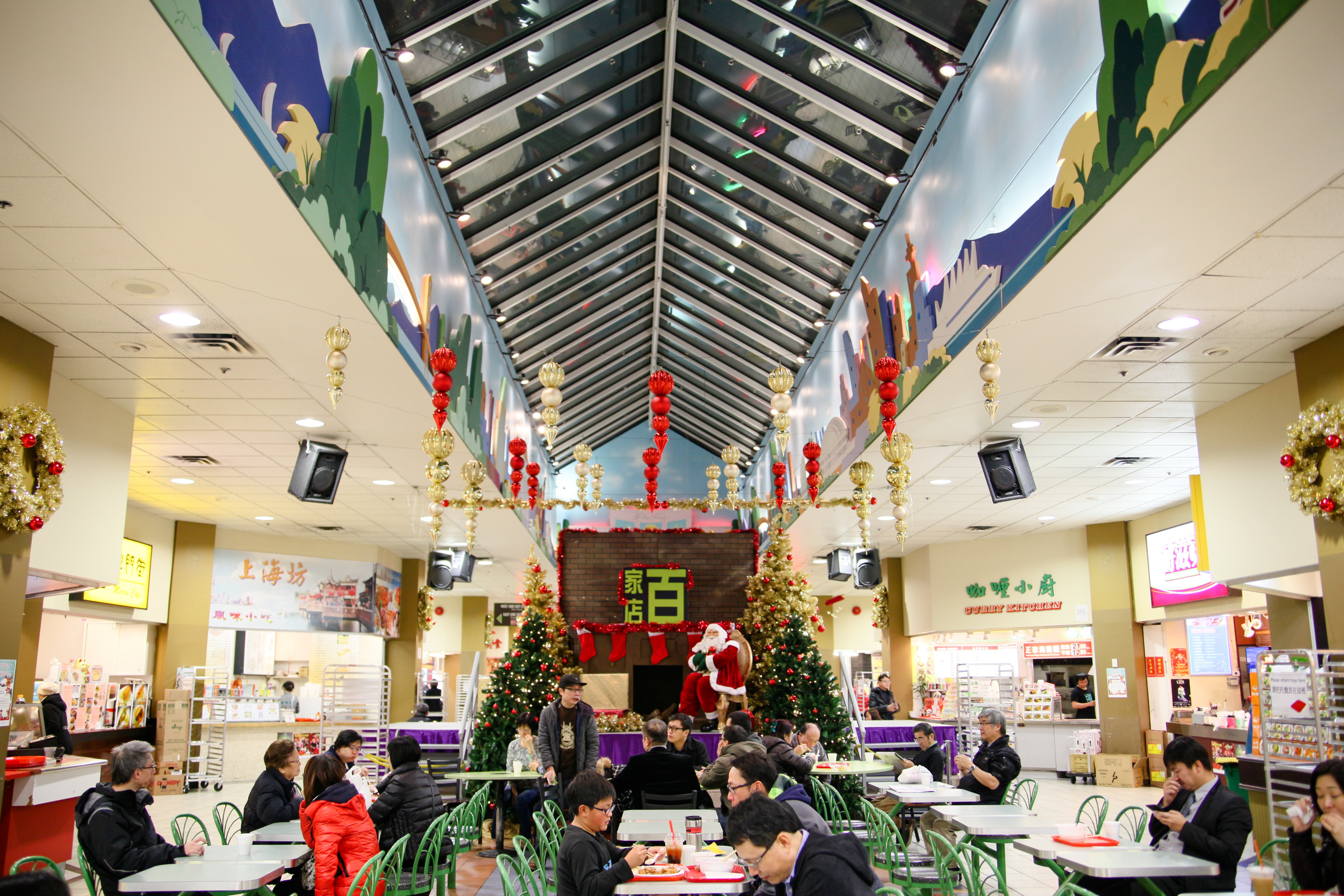 A food court at Parker Place Mall in Richmond, BC — one of the region’s first Chinese malls — is decorated for Christmas with gold wreaths, gold and red icicle baubles and Santa Claus on a purple riser in the background. In the foreground are people seated at tables, resting or eating lunch.