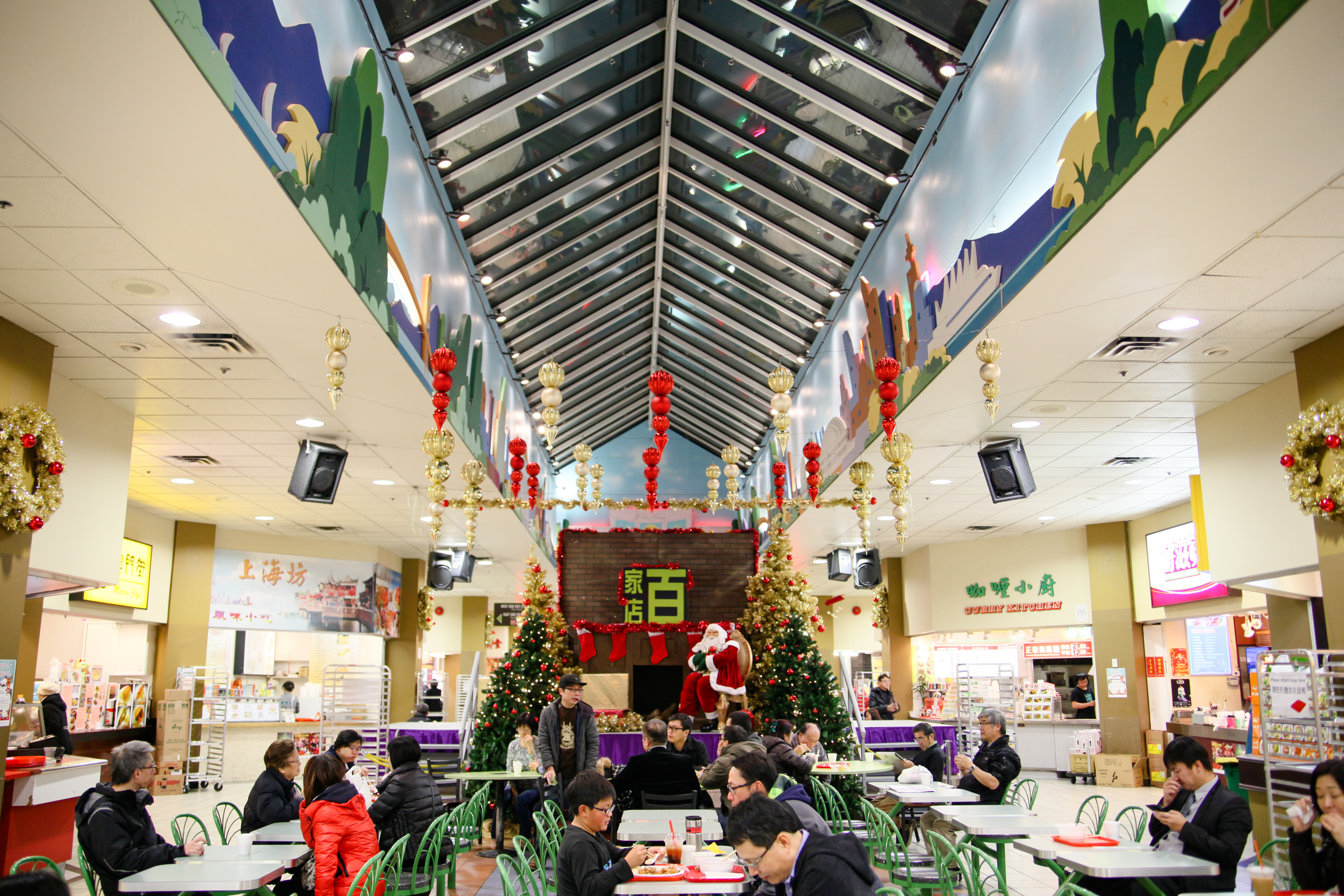 A food court at Parker Place Mall in Richmond, BC — one of the region’s first Chinese malls — is decorated for Christmas with gold wreaths, gold and red icicle baubles and Santa Claus on a purple riser in the background. In the foreground are people seated at tables, resting or eating lunch.