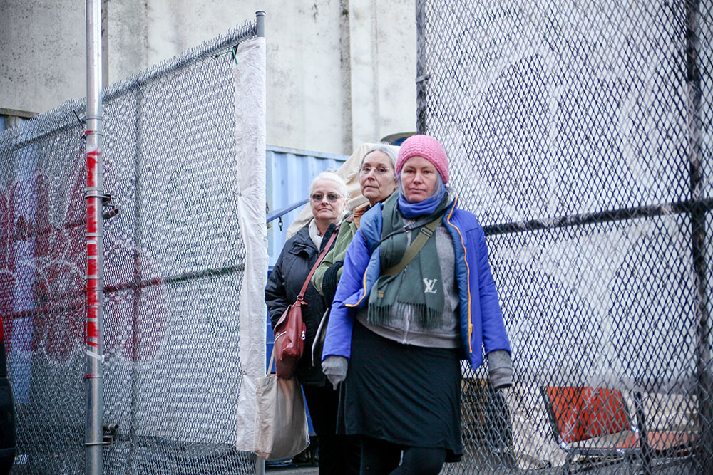 Three women stand together against chain-link fences in front of a grey concrete wall and light blue shipping container that house the fledgling Overdose Prevention Society in Vancouver’s Downtown Eastside in 2016. Sarah Blyth is wearing a pink toque and blue jacket. Behind her are Overdose Prevention Society co-founders Ann Livingston (in a green jacket) and Chris Ewart (in dark glasses).