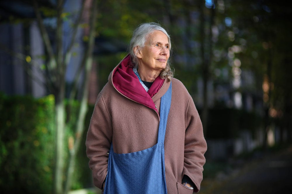 A woman with grey hair in a low ponytail gazes to her left. She is wearing a light pink coat with a big hood. Townhomes are blurry but visible in the background, on a leafy street.