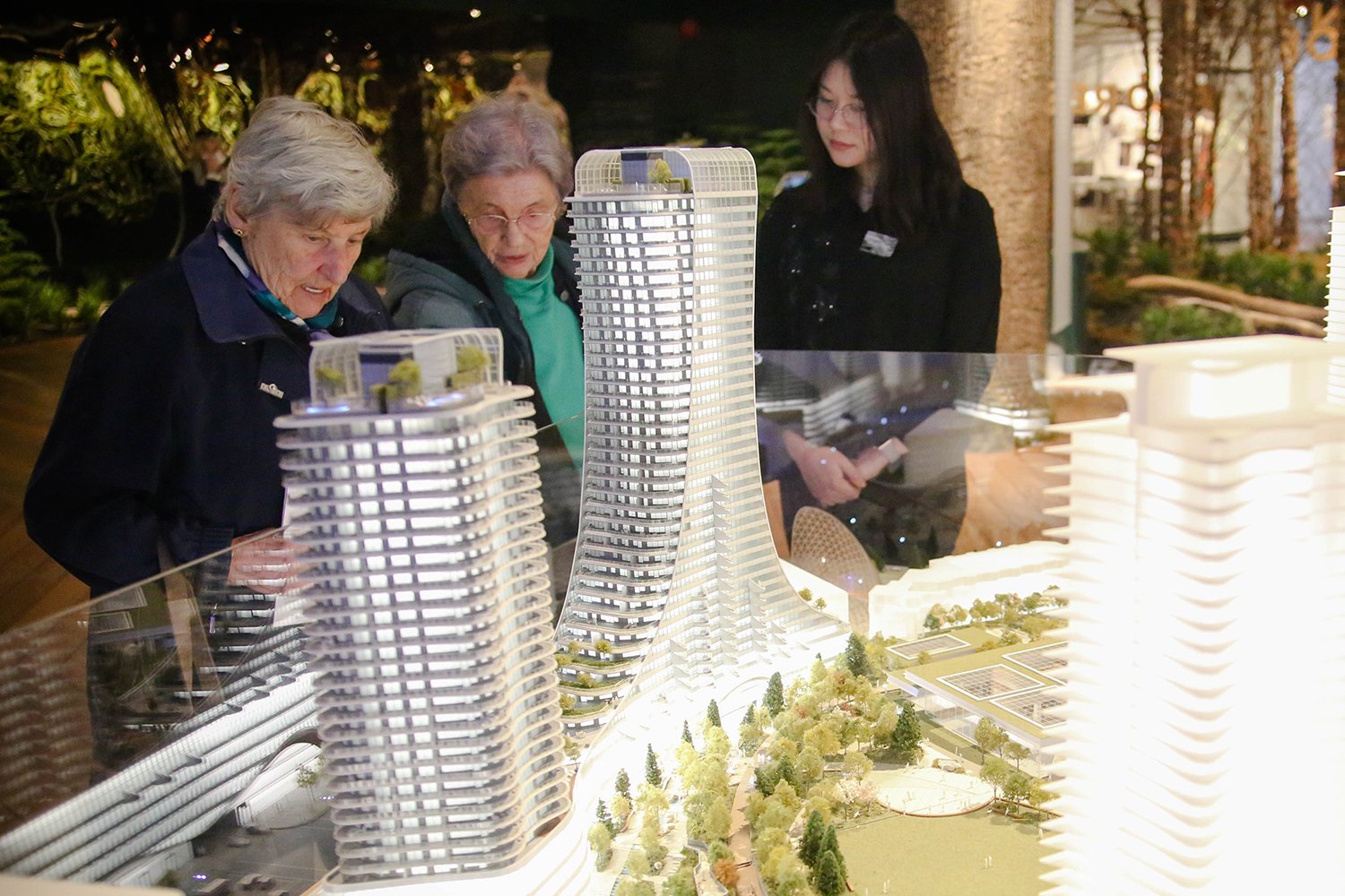 Two elderly women chat in front of a glowing architectural model of a large development’s curvy towers.