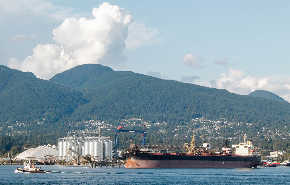 A wide-angle picture shows a large tanker parked in front of industrial lands with mountains in the background. 