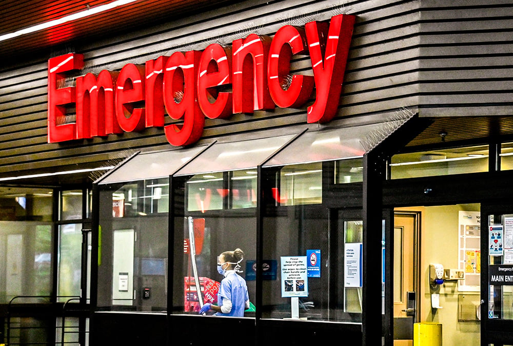 A masked health worker is seen inside hospital windows under a large ‘Emergency’ sign.