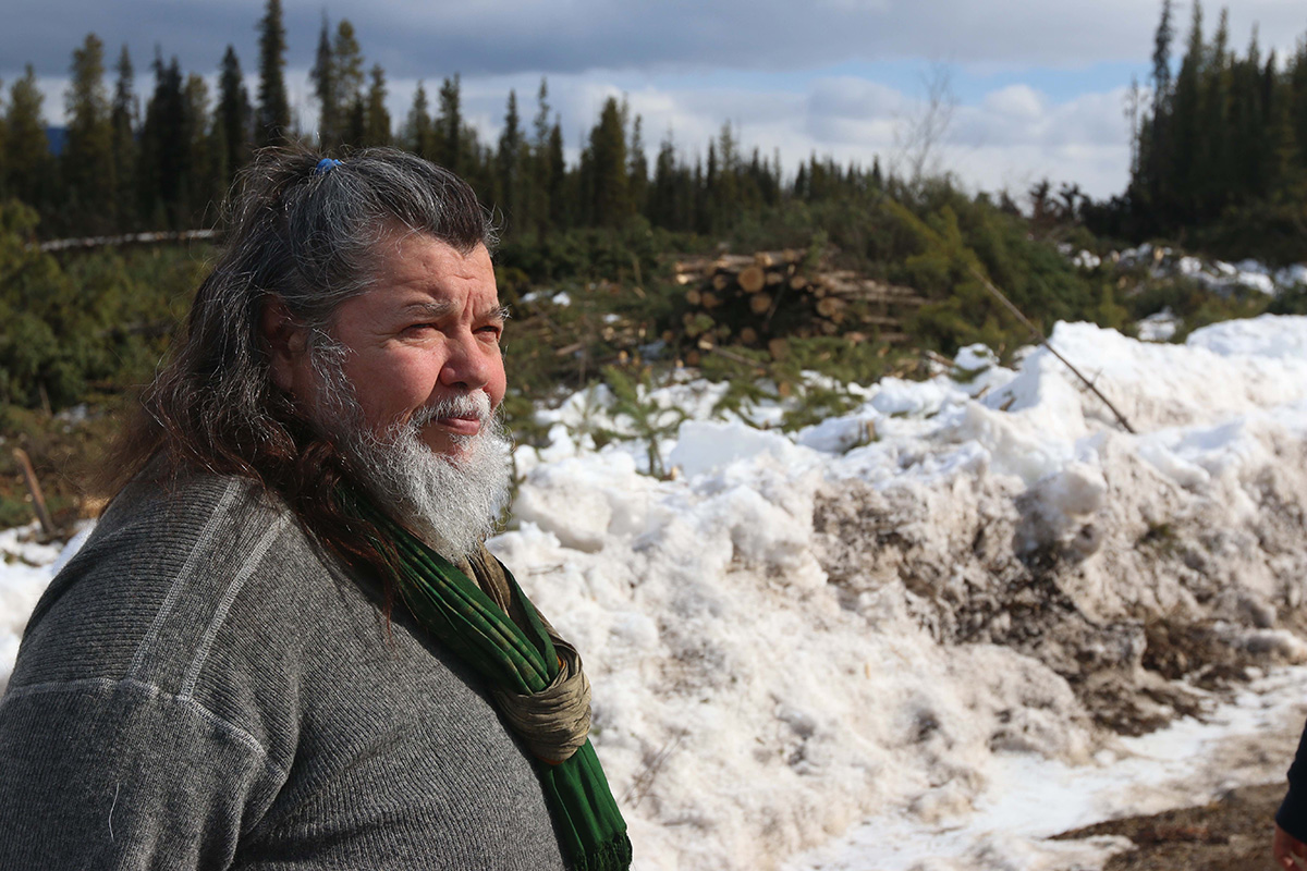 A burly man with long dark hair streaked with grey and white beard standing against a snowy slope.