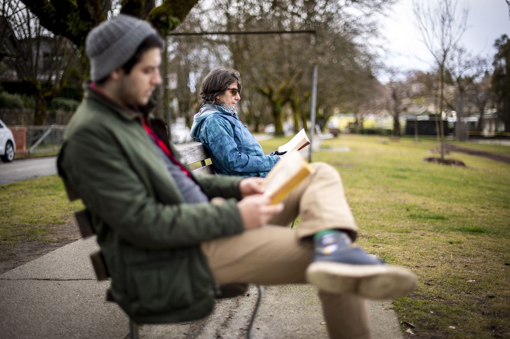An out of focus young man wearing a toque sits reading on park bench, also occupied by a middle-aged woman who is also reading.