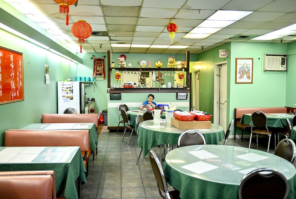 A restaurant dining room with green walls, red banquets and tables with green tablecloths has red decorative Chinese lanterns hanging from the ceiling and an assortment of happy cats and Chinese figurines arranged on the bar and its shelves at the back of the room. Against the bar, a woman with short dark hair and glasses is seated at a table, reading.